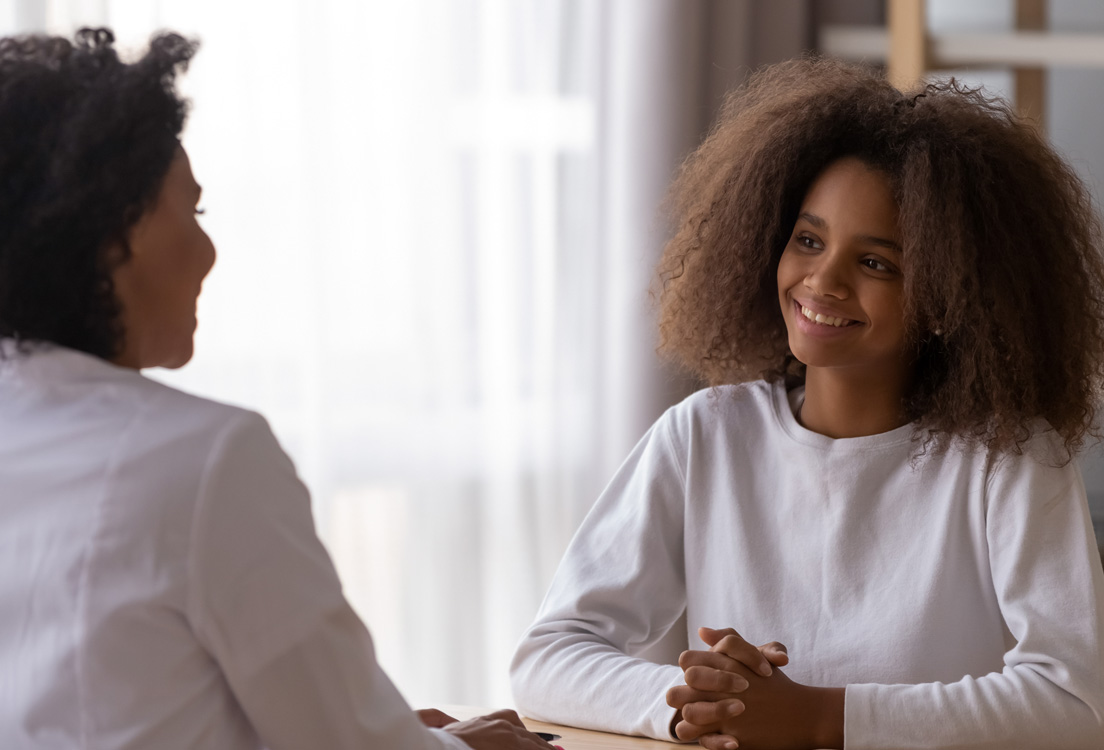 Smiling,African,American,Teenage,Girl,Sit,At,Table,Talking,With Smiling African American teenage girl sit at table talking with female teacher or tutor at home, happy teenager have casual conversation with woman doctor or specialist. Adult children relationships