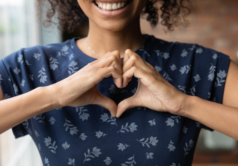 Crop close up of happy African American woman feel grateful thankful show heart sign spread love and care. Smiling biracial female volunteer make hand gesture support ill sick people patients.